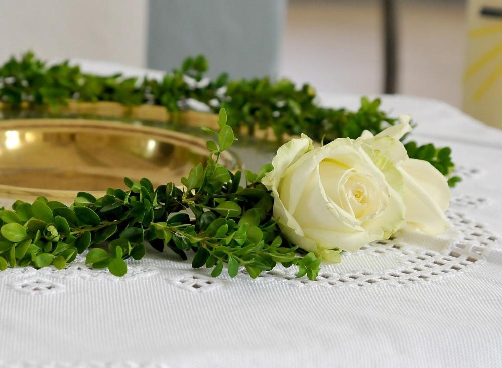 Pöydällä kastemalja, jonka ympärillä vihreitä kasvin lehtiä sekä valkoinen ruusu.