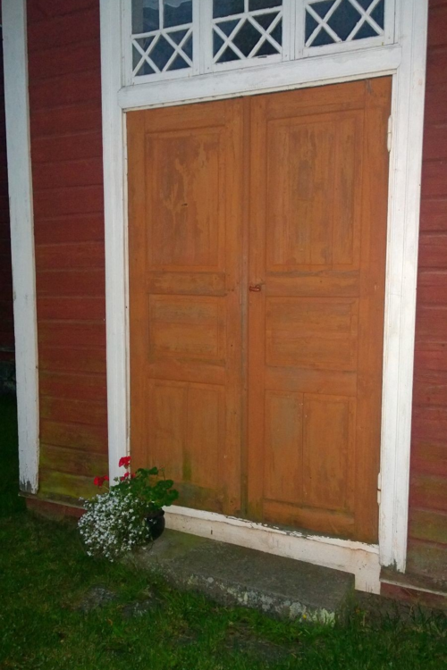 Vanhemman rakennuksen puinen  pariulko-ovi, jonka kivirapulla kukkiva kukka ruukussa.
