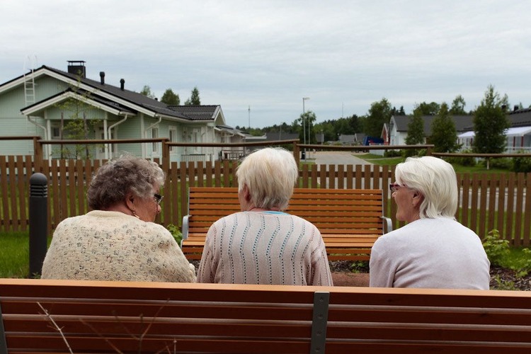 Kolme ikääntynyttä naista istuvat ulkona penkillä juttelemassa selät kameraan päin.